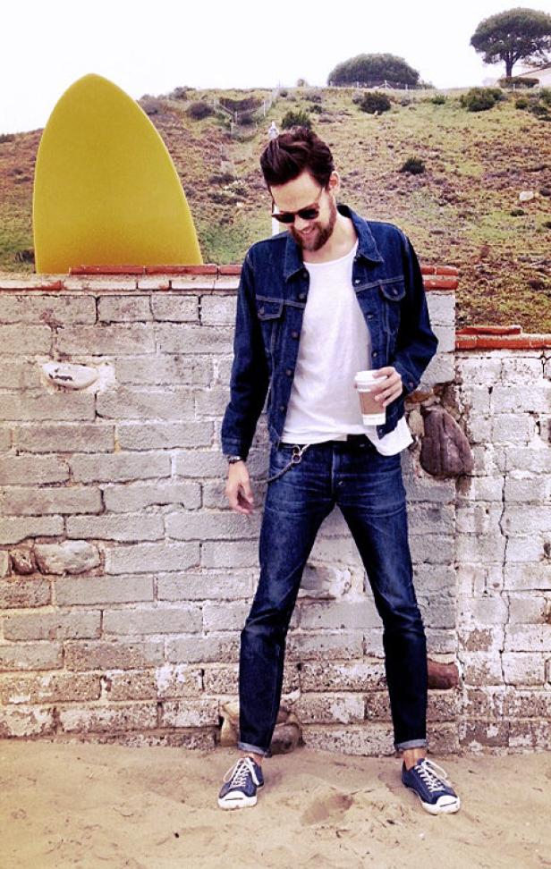 Ein Mann mit Sonnenbrille, Jeansjacke und Kaffee steht vor einer Mauer mit Surfbrett im Hintergrund.