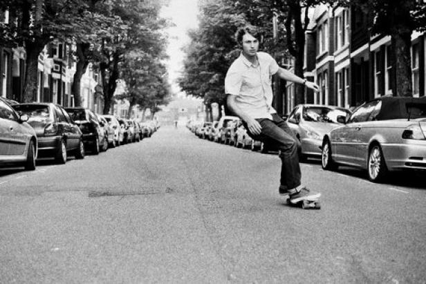 Ein Mann fährt auf einem Skateboard eine von geparkten Autos gesäumte Straße entlang.