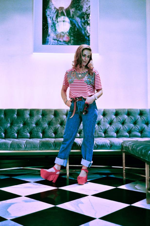 Eine Frau posiert in Jeans, einem gestreiften Oberteil und roten Plateauschuhen vor einem Sofa.