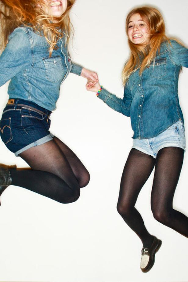 Zwei junge Frauen in Jeansjacken und Shorts springen und halten Händchen.