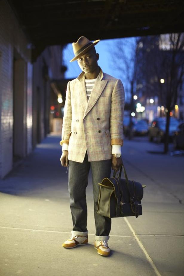 Ein Mann mit Hut, Blazer und Tasche steht auf einem Bürgersteig.