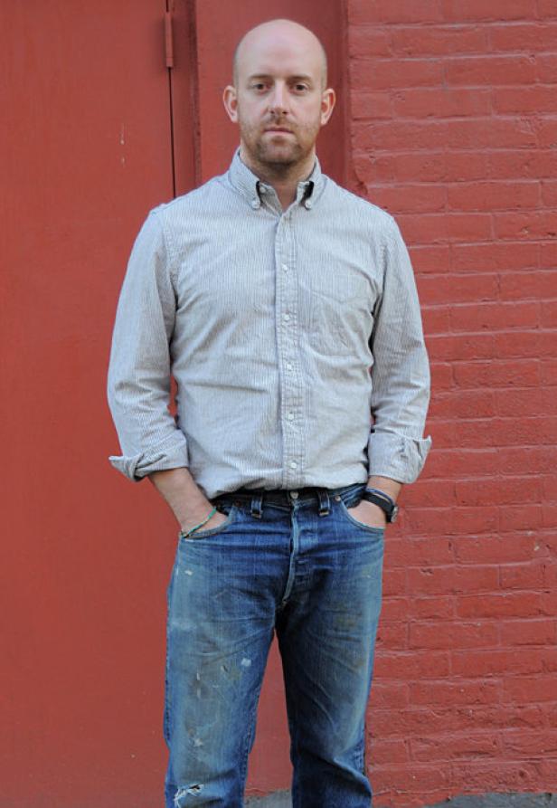 Ein Mann mit Hemd und Jeans steht vor einer roten Backsteinmauer.