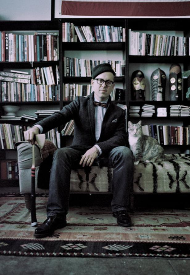 Ein Mann mit Brille und Fliege sitzt mit einer Katze vor einem Bücherregal.