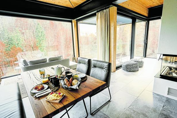 Ein gedeckter Frühstückstisch in einem modernen Haus mit Blick auf einen herbstlichen Wald.