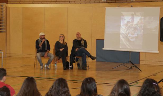 Drei Personen sitzen auf Stühlen vor einer Leinwand, auf der ein Bild von Kindern zu sehen ist.