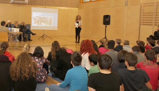 Eine Frau hält einen Vortrag vor einer Gruppe von Schülern in einer Turnhalle. Auf der Leinwand ist ein Bild von Istanbul zu sehen.