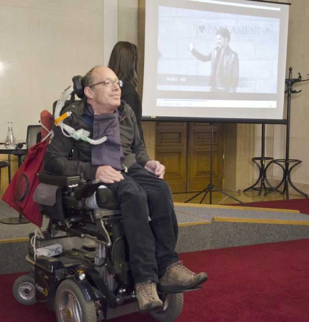 Ein Mann im Rollstuhl sitzt vor einer Leinwand, auf der ein Video abgespielt wird.