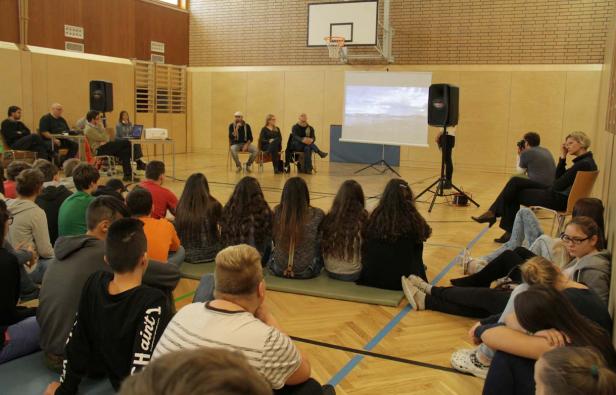 Eine Gruppe von Schülern sitzt in einer Turnhalle und schaut einer Präsentation zu.