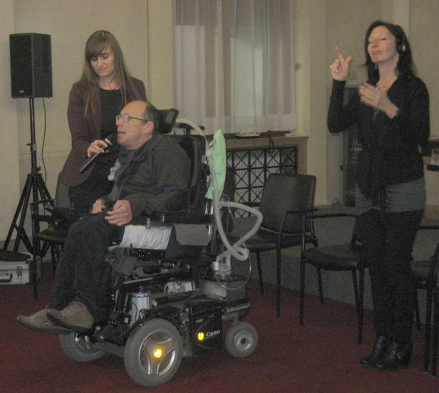 Ein Mann im Rollstuhl spricht mit Mikrofon, während eine Frau in Gebärdensprache übersetzt.