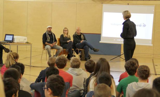 Eine Frau hält vor Schülern einen Vortrag, während drei Personen auf der Bühne sitzen.