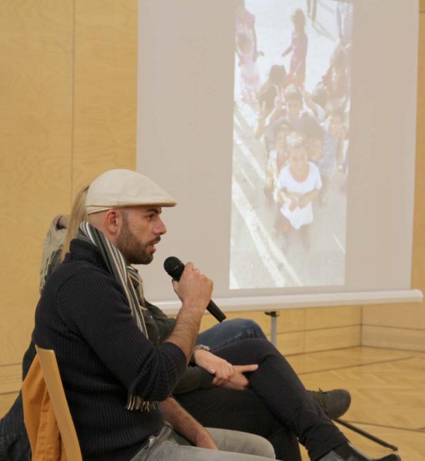 Ein Mann mit Schirmmütze spricht vor einer Projektion von spielenden Kindern.