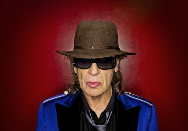 Udo Lindenberg mit Hut und Sonnenbrille vor rotem Hintergrund.