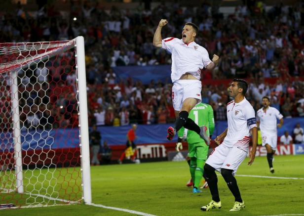 Ein Fußballspieler jubelt auf dem Spielfeld, während seine Mannschaftskameraden ihn feiern.