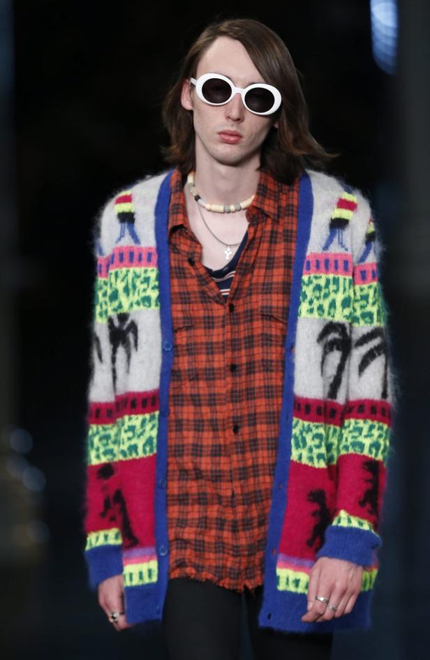 Ein Mann mit Sonnenbrille und buntem Cardigan steht vor einem dunklen Hintergrund.