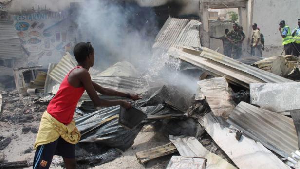 Eine Person gießt Wasser auf die Trümmer eines zerstörten Gebäudes, während im Hintergrund Rauch aufsteigt.
