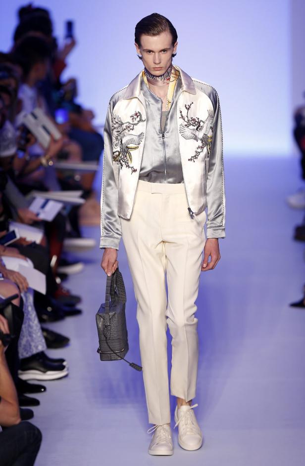 Ein schlanker Mann auf dem Laufsteg präsentiert eine bestickte Jacke und eine graue Handtasche.