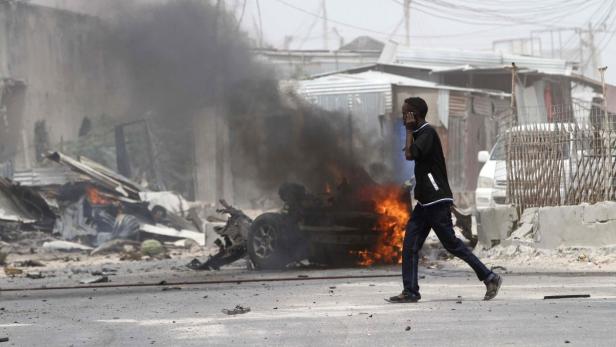 Ein Mann telefoniert, während im Hintergrund ein Auto brennt und Rauch aufsteigt.