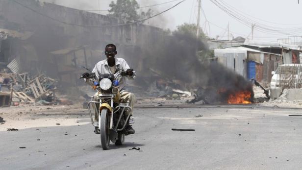 Ein Mann fährt auf einem Motorrad durch eine zerstörte Straße, im Hintergrund brennt ein Feuer.