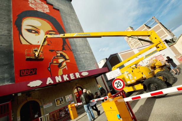 Ein Arbeiter in einem Hubsteiger malt ein großes Wandbild mit dem Schriftzug „Anker“.