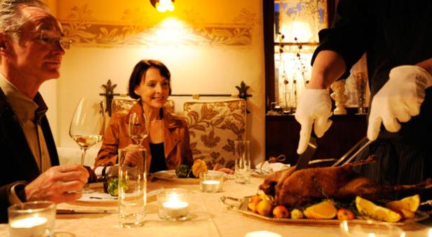 Ein Paar sitzt an einem Tisch, während ein Kellner in weißen Handschuhen eine Gans tranchiert.