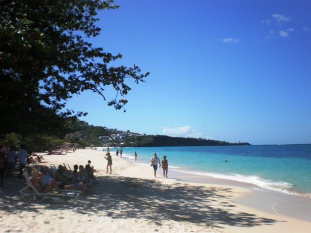 Ein sonniger Strand mit Menschen, türkisfarbenem Wasser und blauem Himmel.