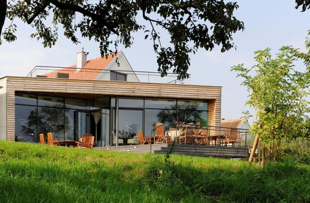 Modernes Haus mit großen Glasfronten, Holzverkleidung und Terrasse mit Holzmöbeln, umgeben von grünem Rasen und Bäumen.