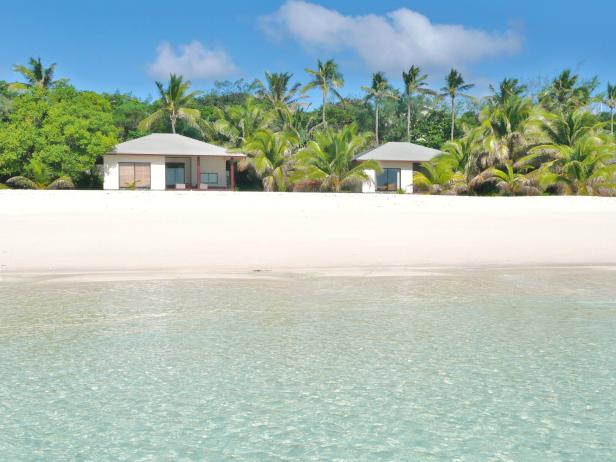 Zwei kleine Häuser stehen an einem tropischen Strand mit Palmen und türkisfarbenem Wasser.