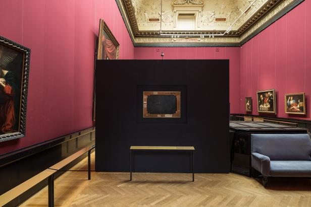 Ein Ausstellungsraum mit Gemälden an weinroten Wänden und einer schwarzen Stellwand.