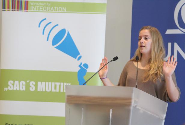 Eine Frau hält eine Präsentation hinter einem Rednerpult mit dem Slogan „Sag’s Multi!“.