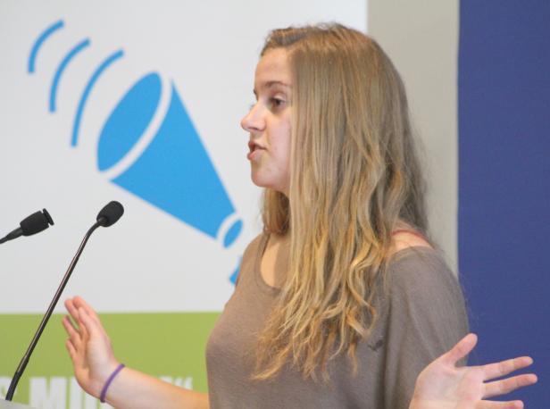 Eine Frau spricht am Rednerpult vor einem blauen Logo mit Mikrofonen.