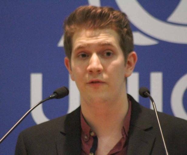 Ein Mann spricht vor einem blauen Hintergrund mit einem Logo an zwei Mikrofonen.