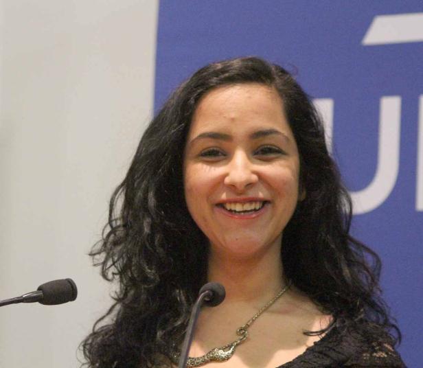 Eine lächelnde Frau spricht an einem Rednerpult mit Mikrofonen.