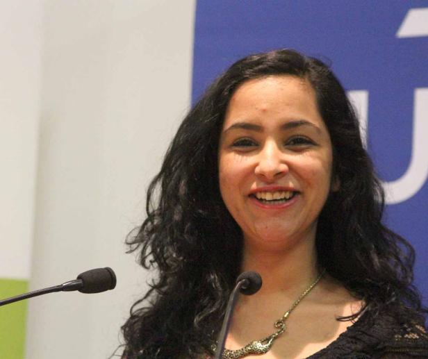 Eine lächelnde Frau mit dunklen, lockigen Haaren steht an einem Rednerpult.