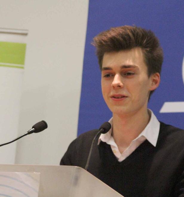 Ein junger Mann spricht an einem Rednerpult mit zwei Mikrofonen.