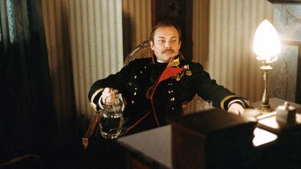 Ein Mann in Uniform sitzt an einem Tisch mit einer Karaffe und einer Lampe.