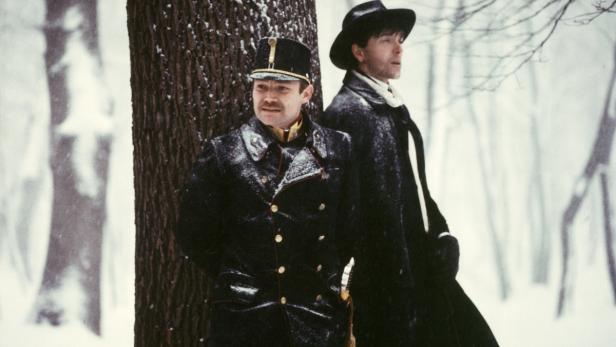 Zwei Männer in Winterkleidung stehen an einem Baum im Schnee.