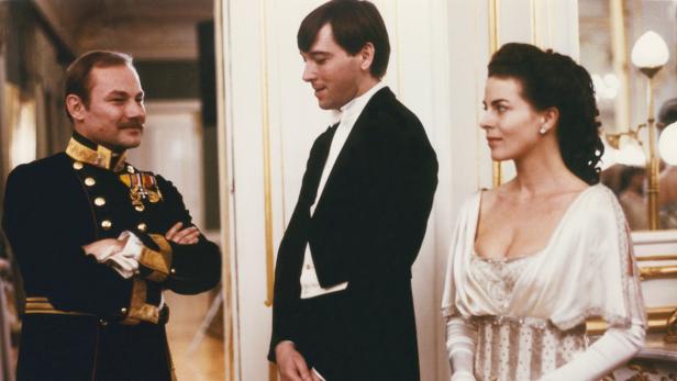 Ein Mann in Uniform, ein Mann im Smoking und eine Frau in einem eleganten Kleid stehen in einem Raum.
