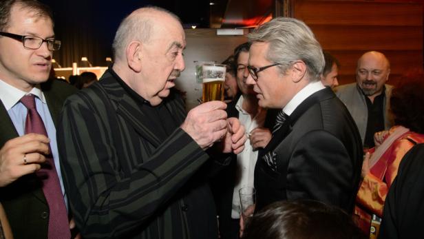 Zwei Männer in Anzügen unterhalten sich, einer hält ein Bierglas hoch.