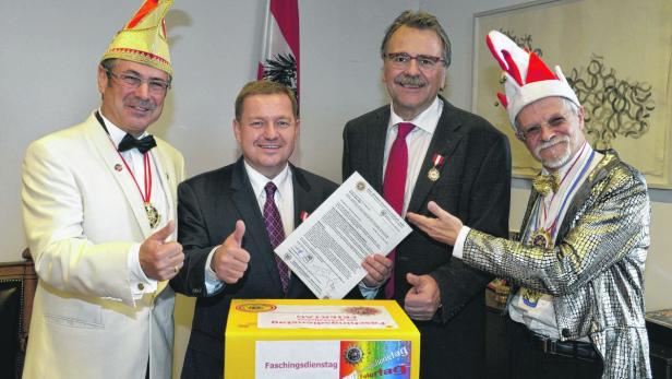 Vier Männer, zwei davon in Karnevalskostümen, posieren mit einer Urkunde und einer gelben Kiste mit der Aufschrift „Faschingsdienstag“.