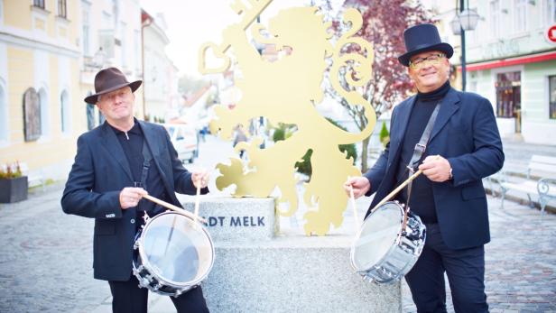 Zwei Männer mit Hüten und Trommeln stehen vor dem Stadtwappen von Melk.