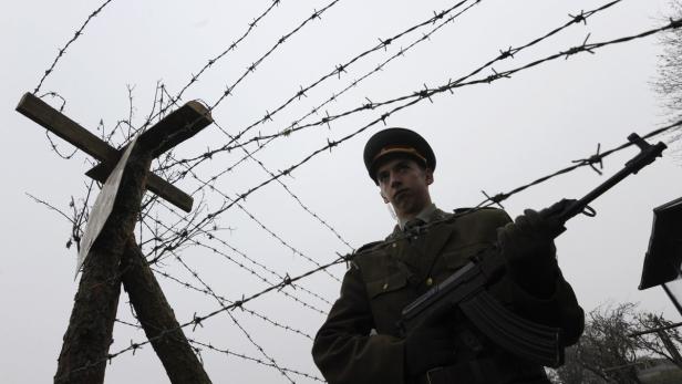 Ein Soldat in Uniform steht mit einem Gewehr vor einem Stacheldrahtzaun.