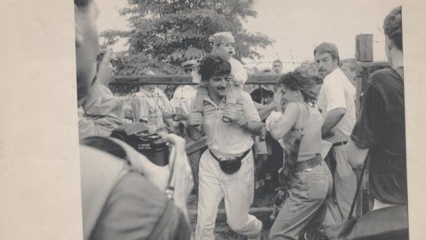 Ein Mann trägt ein Kind auf den Schultern durch eine Menschenmenge hinter einem Zaun.