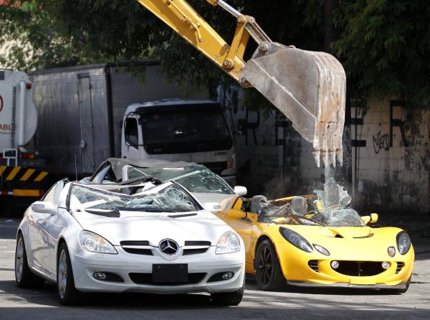 Ein Bagger zerstört einen gelben Lotus und einen weißen Mercedes.