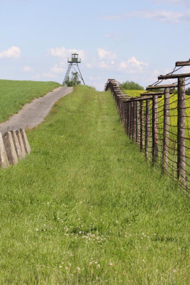 Ein Grenzzaun mit Stacheldraht und ein Wachturm in einer grünen Landschaft.
