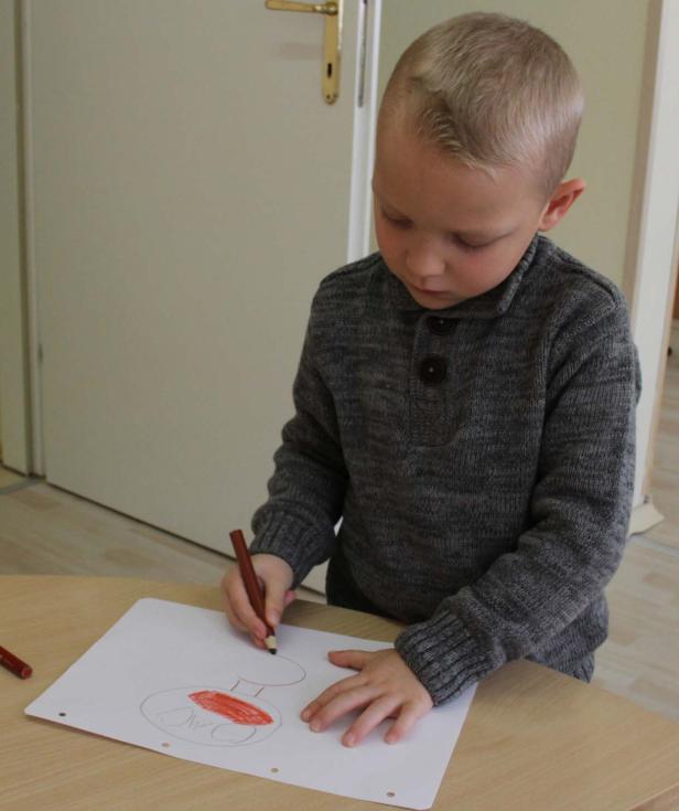 Ein Junge mit grauem Pullover malt mit einem Farbstift auf einem Blatt Papier.