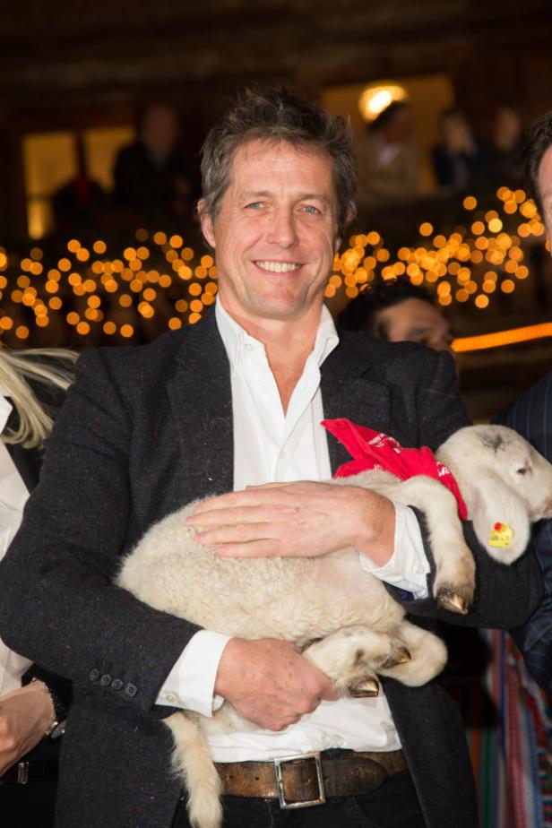 Hugh Grant hält ein Lamm mit rotem Halstuch im Arm.