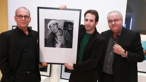 Drei Männer präsentieren ein gerahmtes Schwarzweißfoto eines älteren Mannes mit einer Puppe.