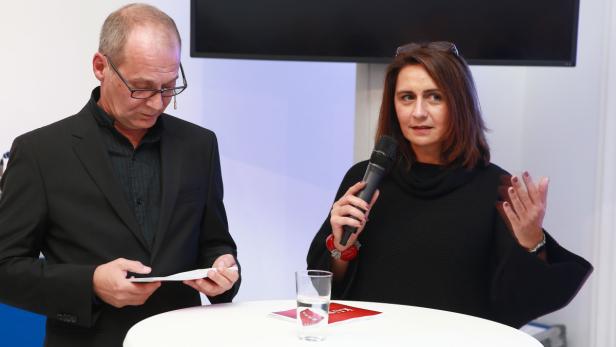 Eine Frau spricht mit einem Mikrofon, während ein Mann daneben in ein Dokument schaut.
