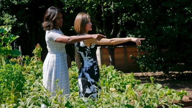 Michelle Obama und Königin Letizia von Spanien zeigen in einem Garten in die Ferne.