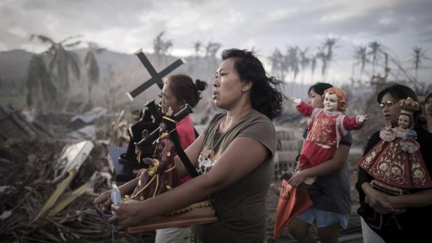 Frauen tragen religiöse Statuen durch eine zerstörte Landschaft.